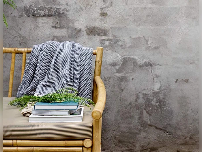 En bambusbænk med en stribet hynde, et foldet gråt tæppe og en stak bøger med grønt løv på toppen står op ad en struktureret betonvæg.