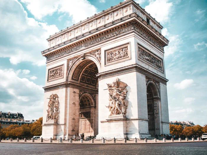 Triumfbuen står højt under en delvist overskyet himmel med detaljerede udskæringer på facaden og en brostensbelagt gade i forgrunden.