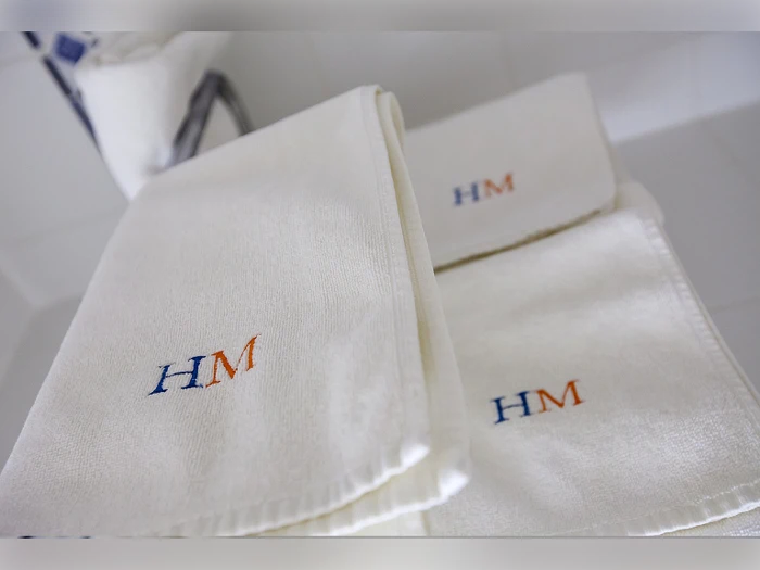 Tre hvide håndklæder med HM broderet i blå og orange tråd ligger pænt foldet på en hvid flade. En toiletrulle er synlig i baggrunden.