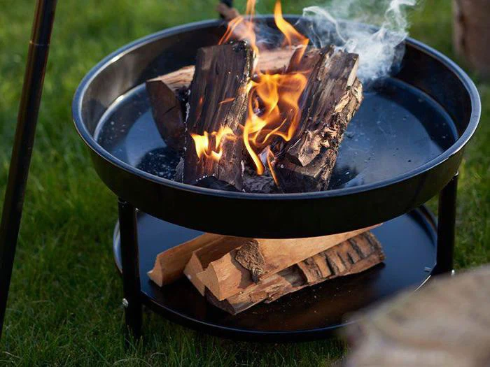 Et sort metalbålsted med brændende træstammer på toppen og utændt brænde på en lavere hylde, placeret på grønt græs udendørs.