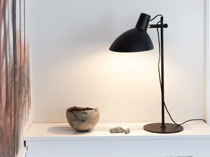 En sort skrivebordslampe oplyser en hvid flade ved siden af en rund, rustik skål og to små sten mod en almindelig hvid væg.