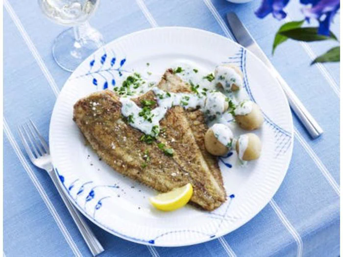 En hvid tallerken med to stykker stegt fisk, små kogte kartofler med flødesauce, hakkede krydderurter og en citronbåd på en blå dug, med gaffel og kniv placeret ved siden af tallerkenen.