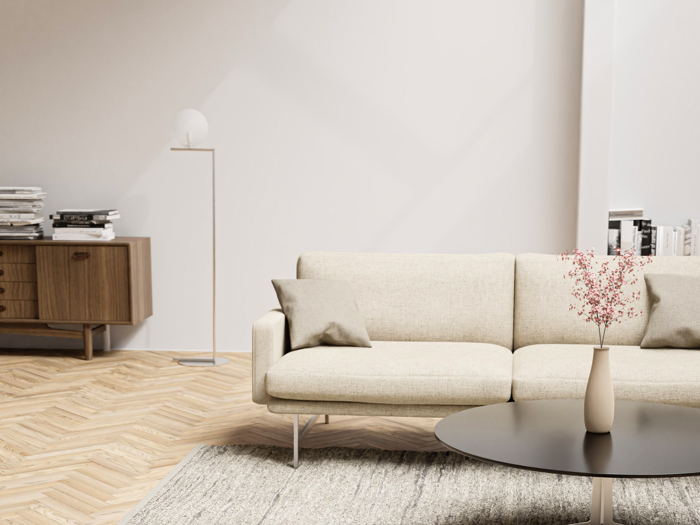 En moderne stue med en beige sofa, to puder, et rundt sort sofabord med en vase med lyserøde blomster, en træskænk og en gulvlampe på et sildebensgulv.