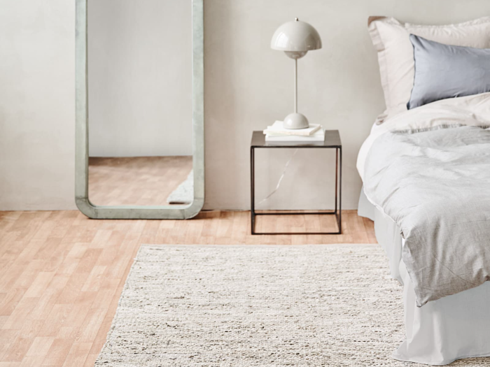 Et minimalt soveværelse med en seng, puder, et natbord med en lampe, en stak bøger, et højt spejl, et lyst tæppe og trægulv.