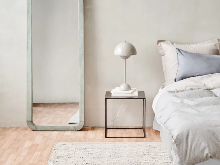 Minimalistisk soveværelse med et stort gulvspejl, moderne natbord, hvid lampe og en pænt redt seng med neutralt sengetøj på et lyst trægulv.