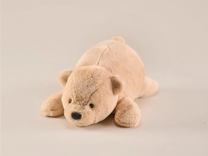 En blød, lysebrun plysbjørn ligger fladt på maven mod en almindelig, lys baggrund.