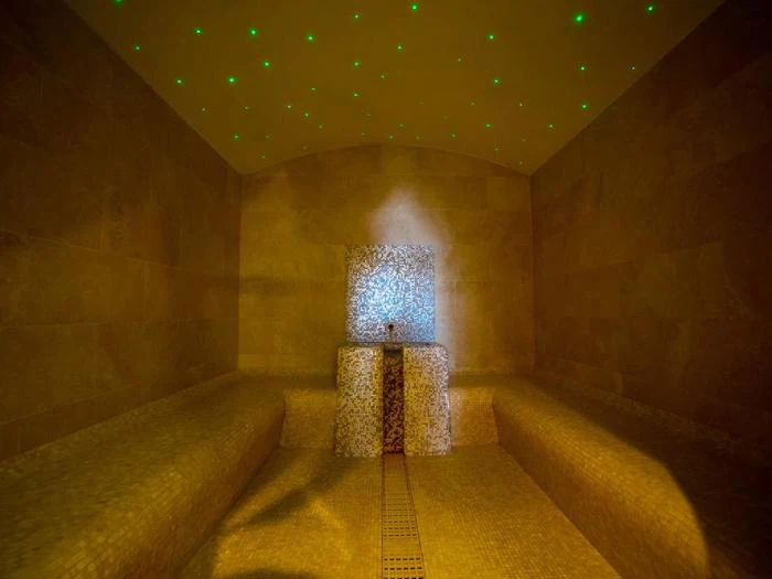 Et flisebelagt dampbad med to bænke, et springvand af mosaikfliser i midten og små grønne lys i loftet, der skaber en stjerneeffekt.