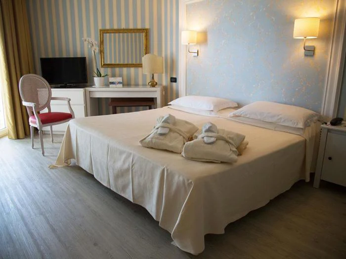 En pænt redt dobbeltseng med to sammenfoldede morgenkåber, et skrivebord med spejl, tv, stol og væglamper i et hyggeligt, lyst hotelværelse med stribet og blomstret tapet.