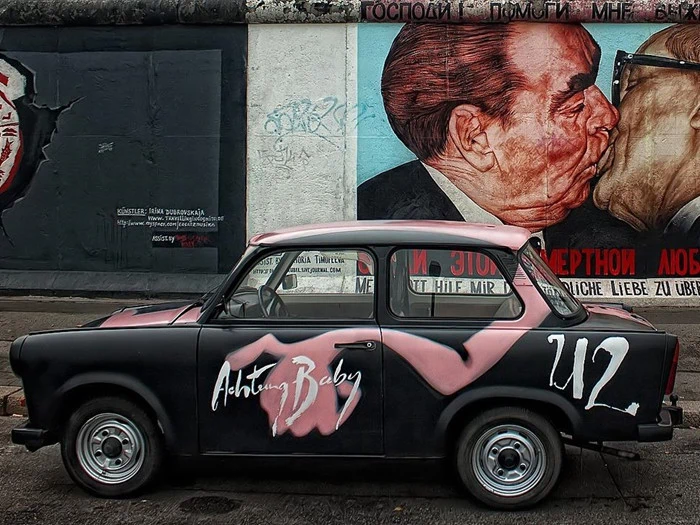 En sort og pink bil med Achtung Baby malet på siden er parkeret foran et vægmaleri af to mænd, der kysser hinanden.