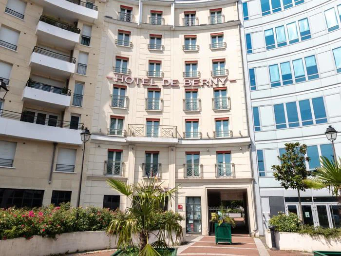 Facaden på Hôtel de Berny, en beige bygning i flere etager med røde vinduer, omgivet af planter og moderne nabobygninger.
