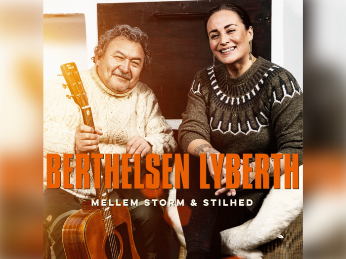 En ældre mand med en guitar og en smilende kvinde sidder sammen, begge iført strikkede trøjer. Tekst: BERTHELSEN LYBERTH og MELLEM STORM & STILHED.