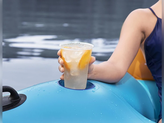 En person i blå badedragt sidder på en blå oppustelig poolflåde og holder en plastikkop med is og citronskiver over vandet.