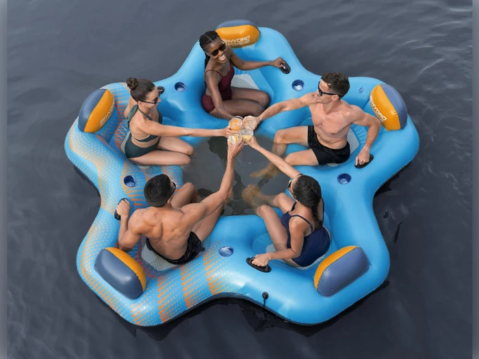 Fire personer i badetøj sidder på en blå oppustelig flyder i vandet, løfter deres drinks sammen i en skål og smiler.