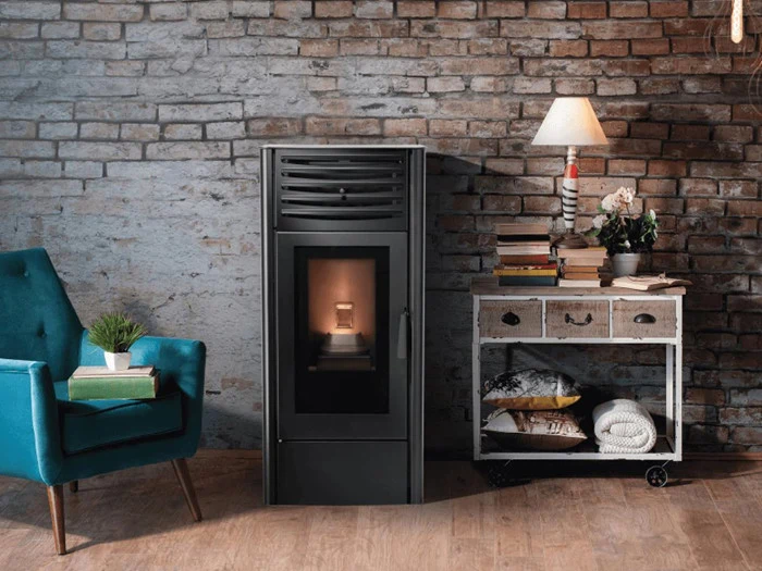 En moderne sort brændeovn står op ad en murstensvæg ved siden af en blågrøn lænestol og et hvidt vintagebord med bøger, en lampe, planter og sammenfoldede tæpper.