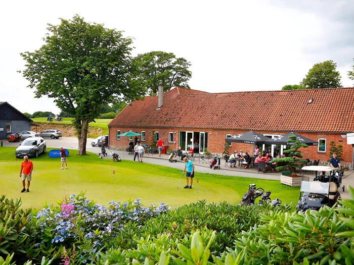 Folk spiller golf og slapper af på en terrasse uden for et klubhus i røde mursten med tegltag, omgivet af grønne områder og golfbags.