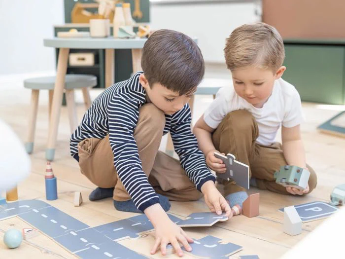 To unge drenge sidder på gulvet og leger med legetøjsbrikker og klodser, mens de bygger en miniatureby sammen i et lyst rum.