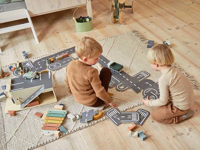 To små børn sidder på et tæppe og leger med legetøjsbiler, træklodser og en vej på et trægulv i et hyggeligt værelse.