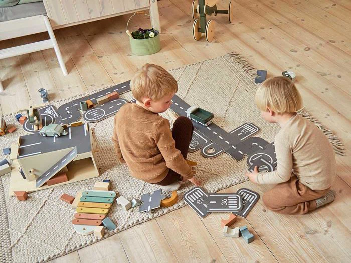 To små børn leger på et tæppe med legetøjsbiler og et vejsæt af pap, omgivet af træklodser og legetøj i et hyggeligt, lyst rum.