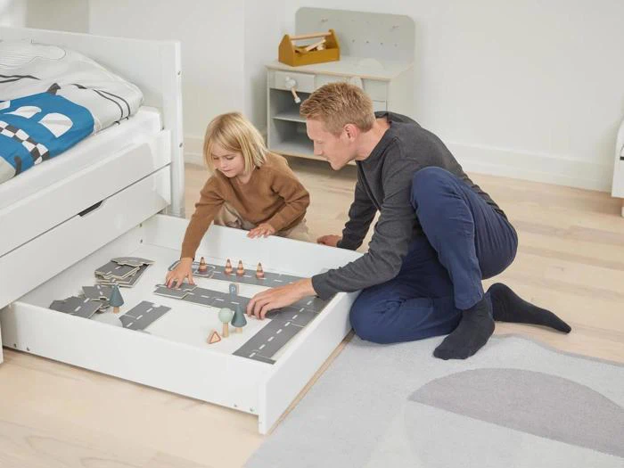 En mand og et lille barn sidder på gulvet og leger med en legetøjsvej og miniaturefigurer i en udtræksskuffe under en seng i et lyst, ryddeligt værelse.