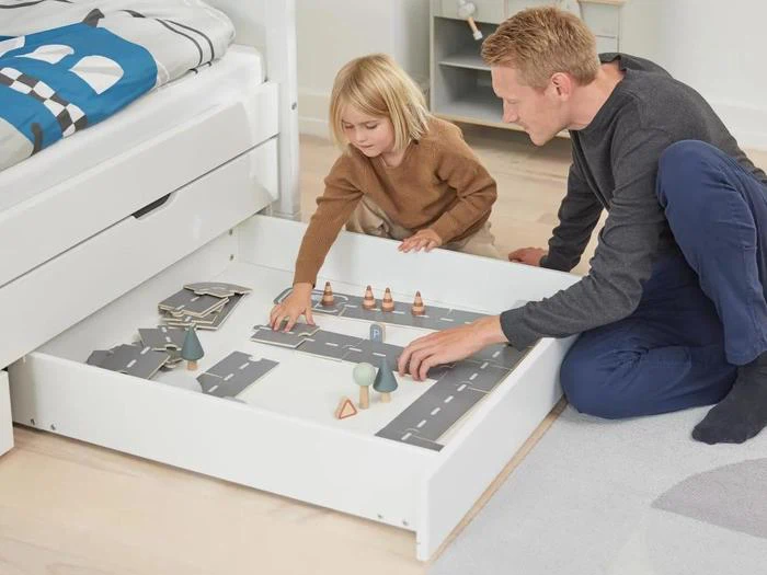 En voksen og et barn leger med en legetøjsvej og køretøjer, der ligger i en udtræksskuffe under en seng i et lyst soveværelse.