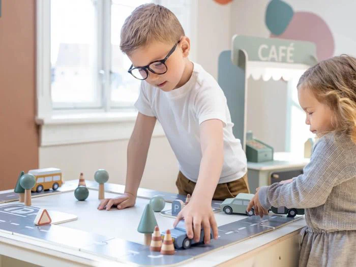To små børn leger med legetøjsbiler og trafikskilte af træ på et bord i et lyst rum med et "CAFE"-skilt i baggrunden.