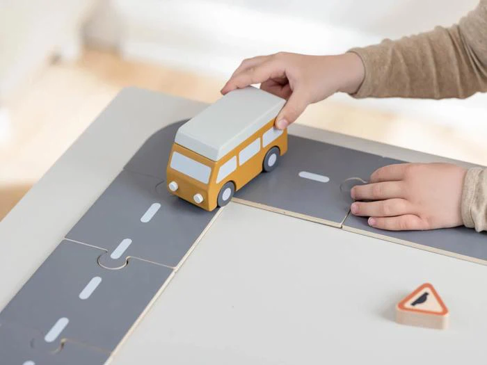 Et barns hænder leger med en gul legetøjsbil på et vejpuslespil på en bordplade med et lille vejskilt i nærheden.