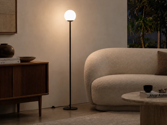 En høj, slank gulvlampe med en lysende, kugleformet pære står mellem et skab af træ og en cremefarvet, buet sofa.