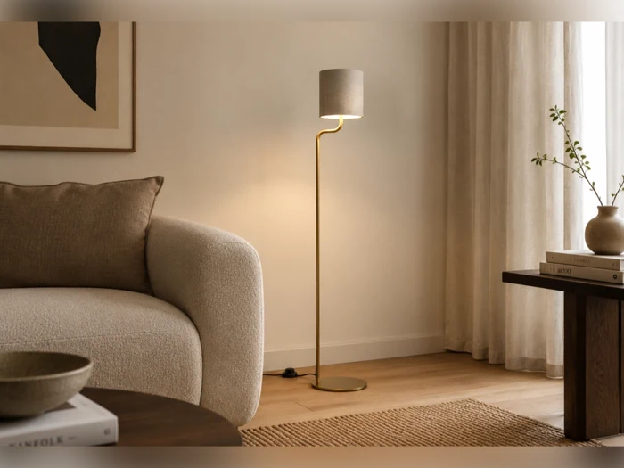 En slank gulvlampe i messing med en lille cylindrisk lampeskærm står mellem en sofa med struktur og et sidebord.