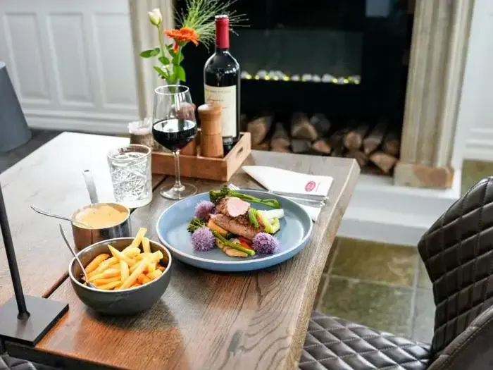 Et træbord med en bøf, pommes frites, et glas rødvin, en flaske vin og et glas vand ved siden af en pejs.