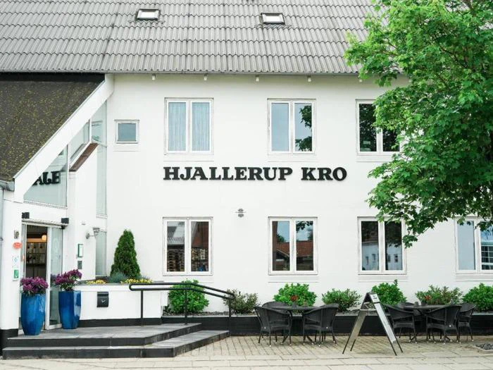 Hvid bygning med HJALLERUP KRO-skilt, udeservering med borde og stole, potteplanter og et træ, der delvist dækker facaden.