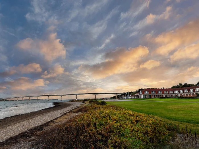 En lang bro strækker sig over vandet nær en stenet kystlinje med en række bygninger med røde tage og en grøn græsplæne under en dramatisk, overskyet himmel ved solnedgang.