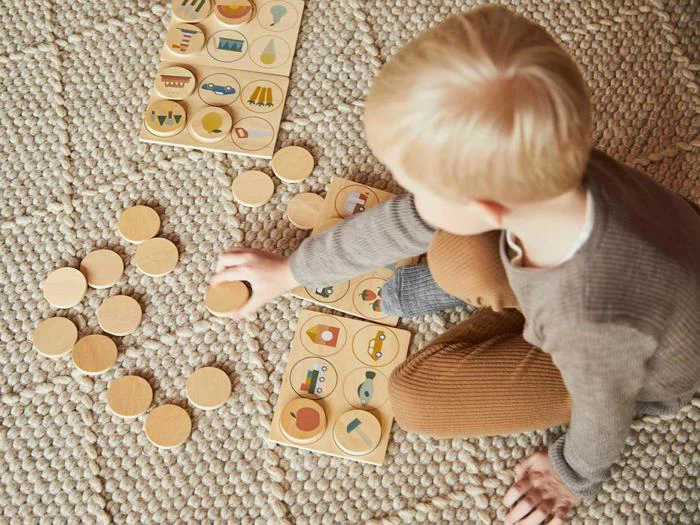 Et lille barn sidder på et tekstureret tæppe og spiller med et matchningsspil i træ med runde brikker og illustrerede plader.