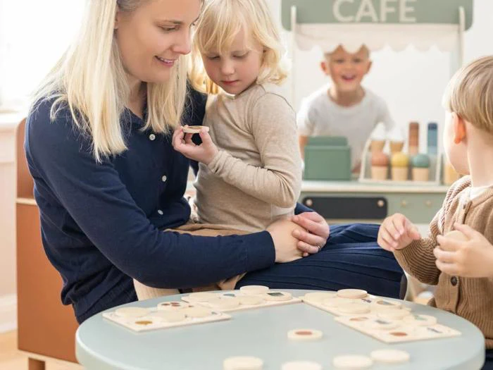 En kvinde og tre små børn leger med træstykker ved et bord i et lyst rum med en legetøjscafé i baggrunden.