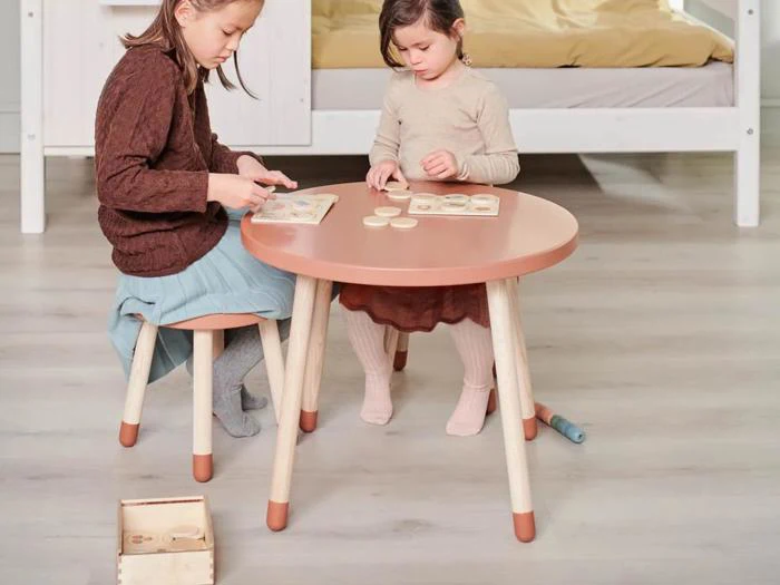 To små børn sidder ved et lille rundt bord og leger med træpuslespil i et hyggeligt, lyst rum med en seng i baggrunden.