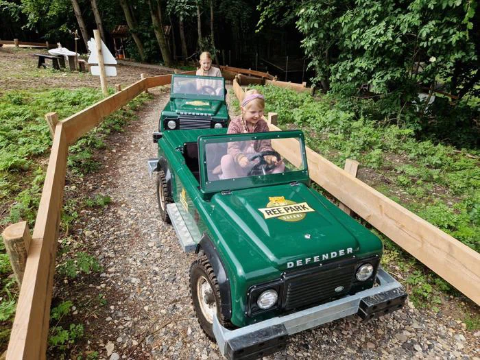 To børn kører i små grønne legetøjsbiler af mærket Land Rover på en grusbane omgivet af grønne områder og træhegn i en udendørs park.