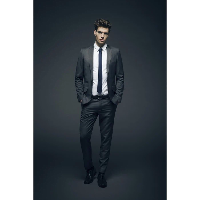 En mand iført gråt jakkesæt, hvid skjorte og sort slips står med hænderne i lommerne mod en mørk baggrund.