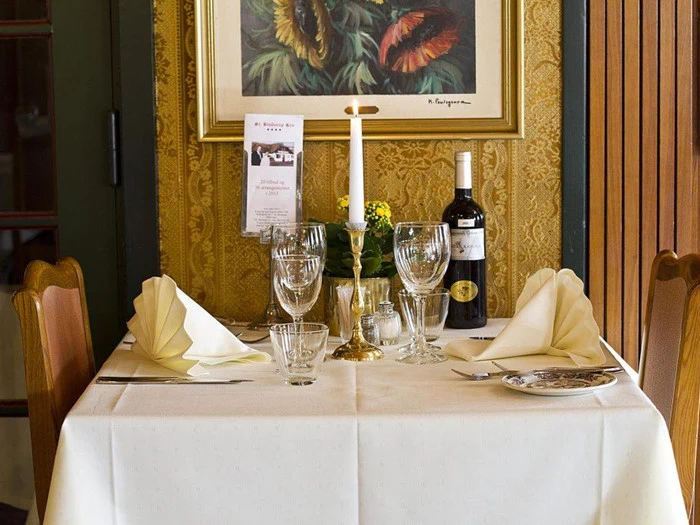 Et bord til to dækket med vin, glas, foldede servietter, et tændt stearinlys og en urtepotte mod en guldmønstret væg med et indrammet maleri.