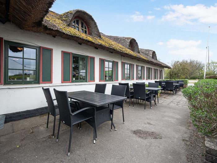 Udendørsservering med sorte borde og stole ved siden af en lang hvid bygning med stråtag og røde vinduer under en blå himmel med spredte skyer.