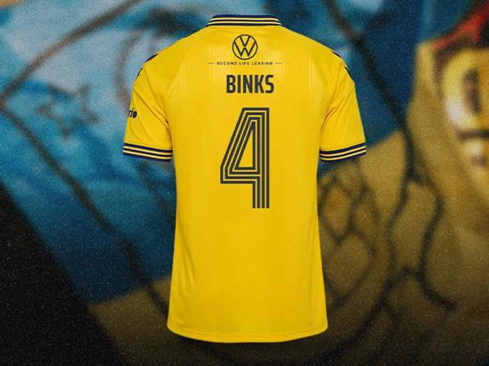 Gul fodboldtrøje med blå detaljer, navnet BINKS, nummer 4 og sponsor NTGI på ryggen, vist mod en stadionbaggrund.