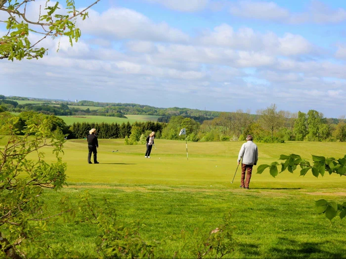 Tre personer spiller golf på en grøn mark under en delvis overskyet himmel med træer og bakker i baggrunden. En person gør klar til at putte, mens de andre ser på.