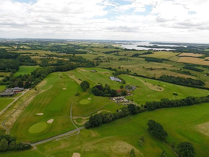 Luftfoto af en golfbane omgivet af grønne marker, træer, en parkeringsplads, bygninger, en dam og fjerne bølgende bakker under en delvist overskyet himmel.