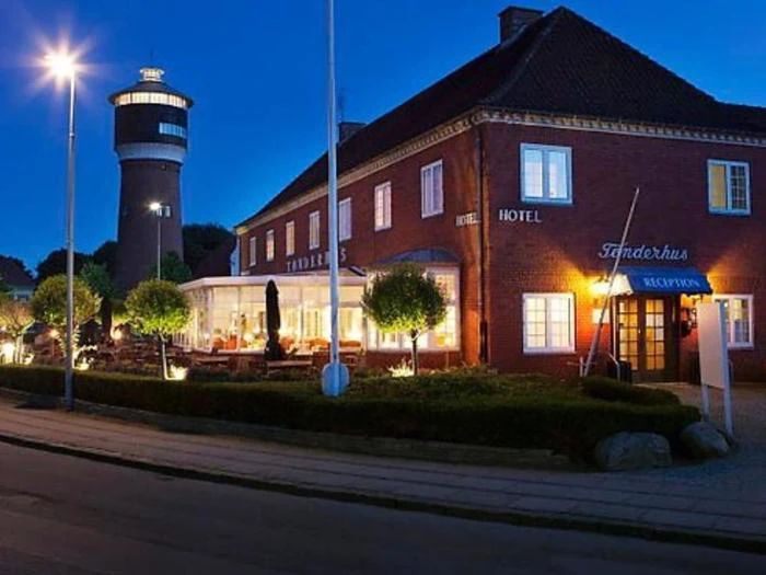 Et murstenshotel med en oplyst glasindgang og udeservering ses i skumringen ved siden af et højt cylindrisk tårn og sirligt klippede buske.