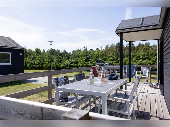 En solrig terrasse med et hvidt bord, stole, drikkevarer og tøj hængt over gelænderet. Frodig grønne omgivelser omkranser de mørke træhuse i baggrunden.