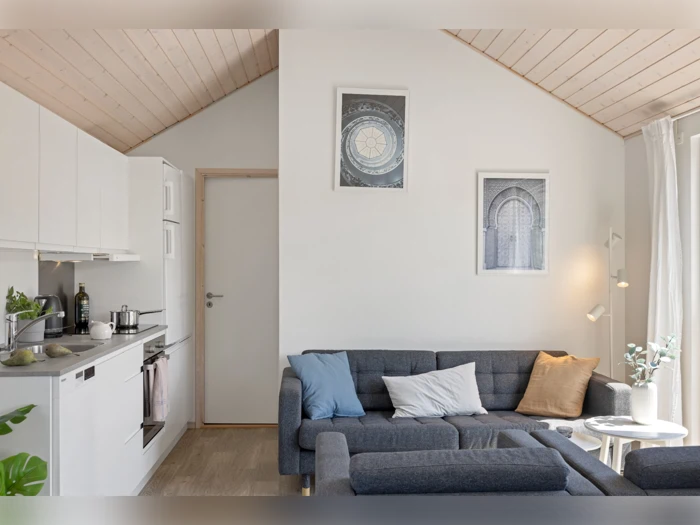 Moderne stue med grå modulsofa, pastelfarvede puder, et lille rundt bord med blomster, hvide vægge, indrammede kunstværker og et køkkenområde til venstre. Loft og gulv i lyst træ.