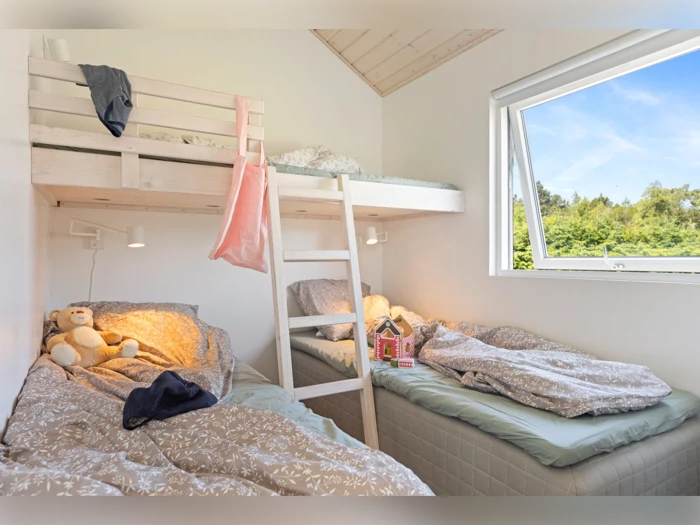 Et hyggeligt soveværelse med en hems, en seng nedenunder, sengetøj, en bamse, et dukkehus og et åbent vindue med udsigt til grønne omgivelser.