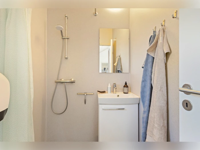 Lille, moderne badeværelse med brusebad, hvid håndvask, spejl, toiletartikler og to håndklæder, der hænger på vægkroge.