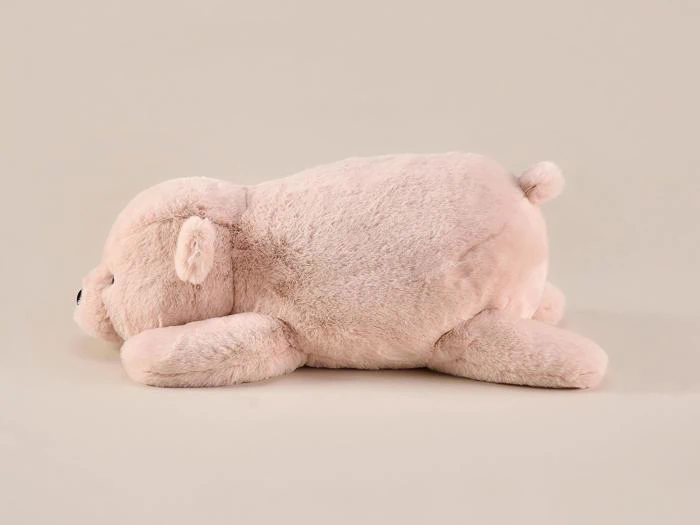 En blød, lyserød plysbjørn ligger på maven mod en almindelig beige baggrund. Bjørnen har en lille hale og lukkede øjne og ser ud til at hvile eller sove.