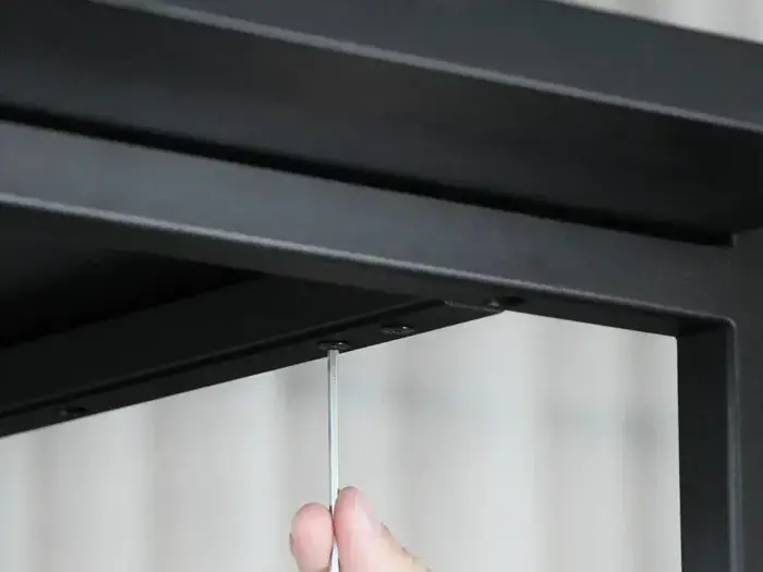 En hånd bruger en unbrakonøgle til at spænde en skrue på en sort metalramme, muligvis ved samling af møbler.