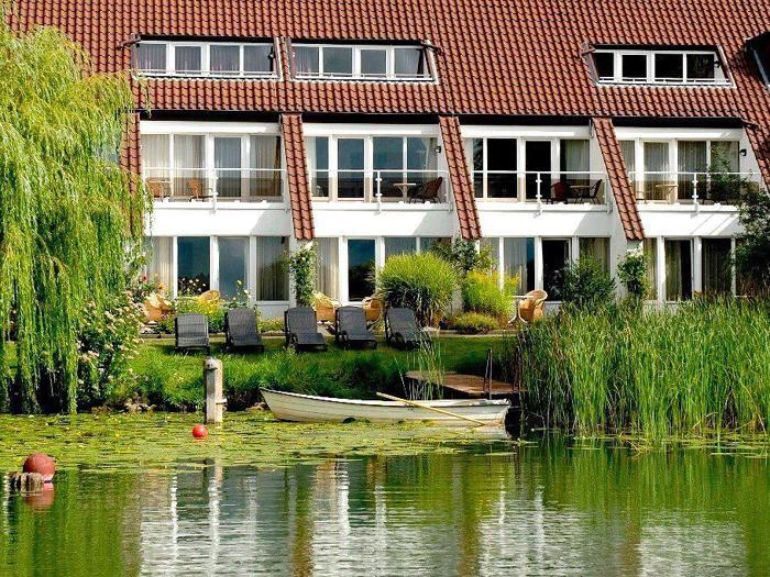 En hvid robåd flyder forbi et hus ved søen med rødt tag, store vinduer, balkoner, liggestole og frodigt grønt, herunder siv og et piletræ.