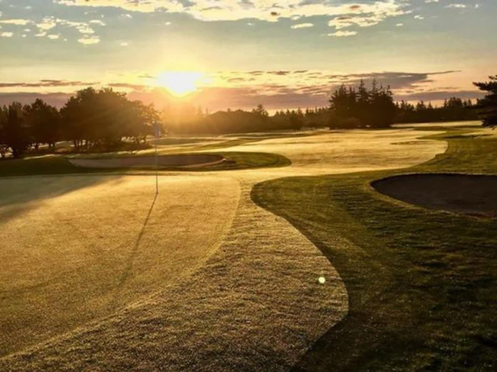Solopgang over en golfbane, der kaster varmt lys og lange skygger over det duggede græs, sandbunkerne og flagstangen i det fjerne.